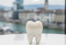 A detailed, close-up photograph of a pristine white ceramic dental crown on a clean, bright surface. The background is softly blurred, revealing architectural elements of the Basel city skyline.
